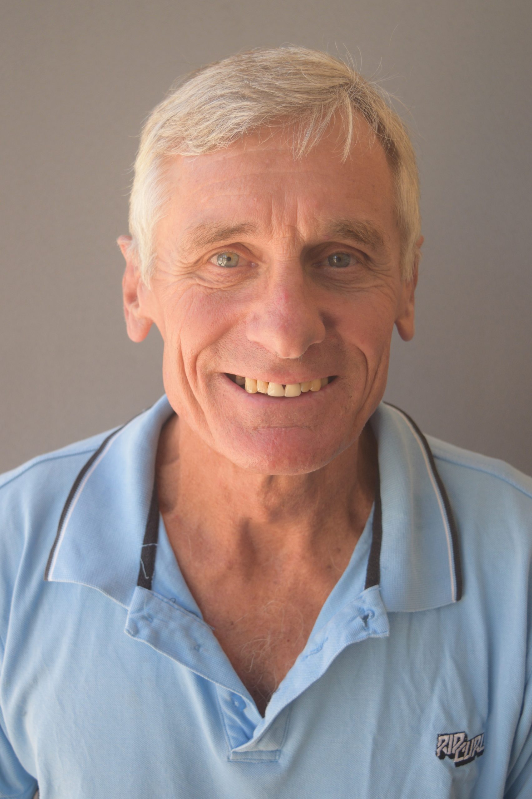 A man with white grey hair, wearing a light blue polo shirt, smiles at the camera
