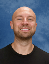 A bald headed man with dark facial stubble smiles at the camera. He's wearing a black t-shirt.